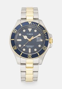 Montre-bracelet en métal argent et or avec un cadran bleu marine, un boîtier rond, des index lumineux et une fenêtre de date à 3 heures. Résistante à l'eau.
