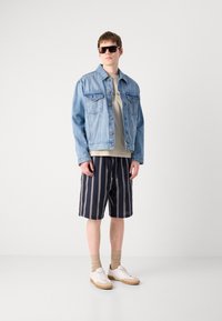 Veste en denim, t-shirt beige, shorts rayés bleu marine, chaussettes beiges et baskets blanches. Les caractéristiques clés incluent une coupe décontractée et des accents bleus sur la veste.