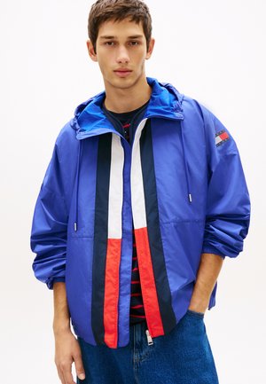 Ung mand iført en blå hættetrøje med røde, hvide og sorte lodrette striber over en sort og rød striped trøje og blå jeans.