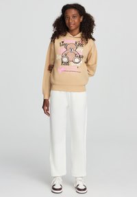 Beżowy hoodie z grafiką misia oraz tekstem w kolorze różowym i czarnym, zestawiony z białymi spodniami o szerokich nogawkach i trampkami z brązowymi dodatkami.