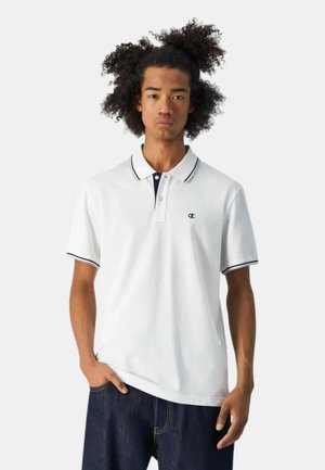 Giovane con capelli ricci che indossa una polo bianca con bordatura nera e jeans blu scuro, in piedi con una mano in tasca.