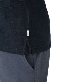 Marineblaues, strukturiertes Poloshirt mit seitlichem Schlitz und weißem Brax-Label, getragen über blaugrauen Hosen mit sichtbarer Tasche.