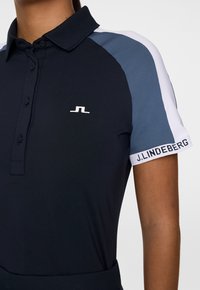 Granatowa koszulka polo z kołnierzem, z jasnoniebieskimi i białymi elementami akcentującymi. W komplecie guziki oraz logo na piersi i rękawie.