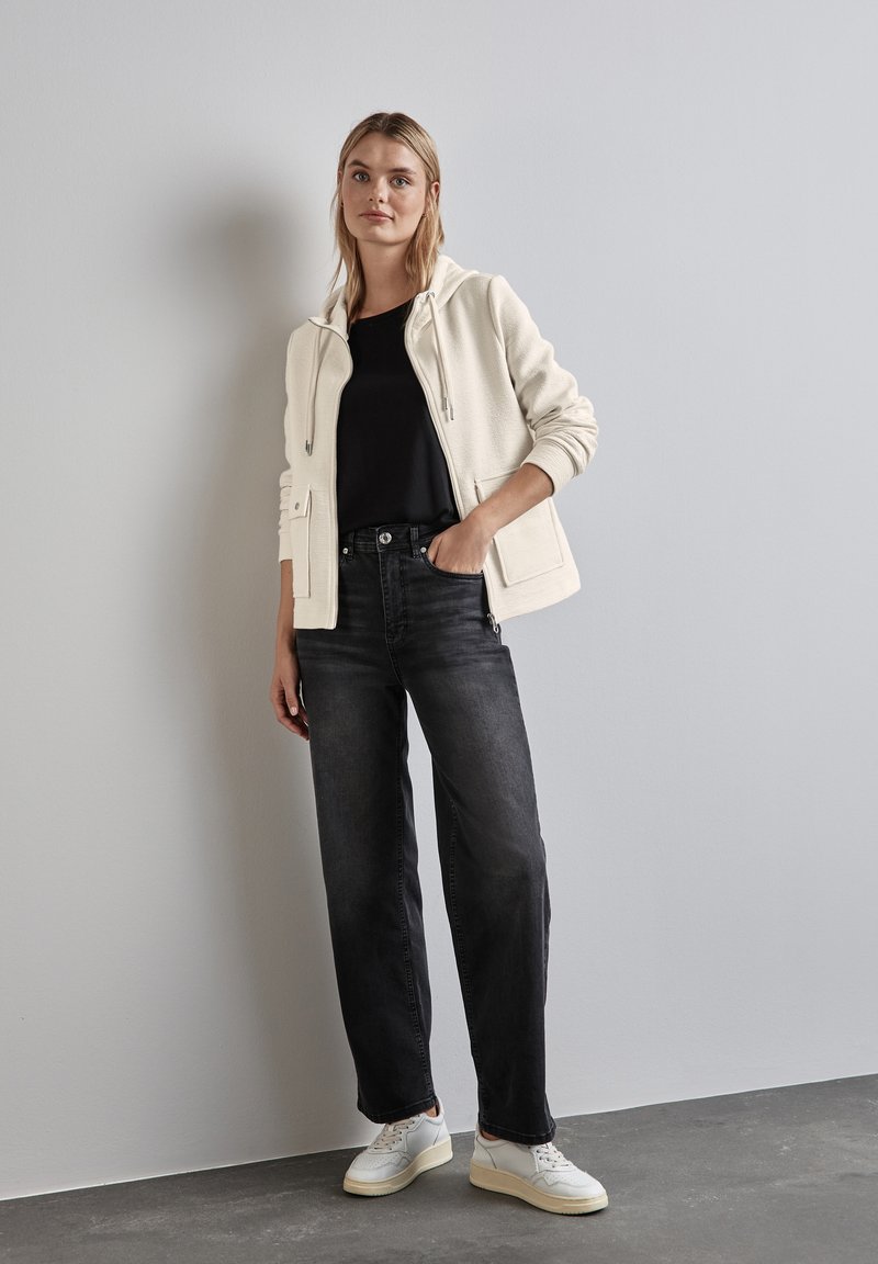 Felpa bianca con zip e tasche frontali su una maglietta nera, abbinata a jeans scuri a vita alta e sneakers bianche, su uno sfondo neutro.