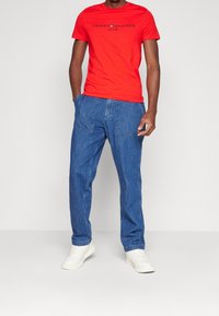 Rød bomulds t-shirt med sort "Tommy Hilfiger" logo, parret med blå denim bukser med to forlommer og hvide sneakers.