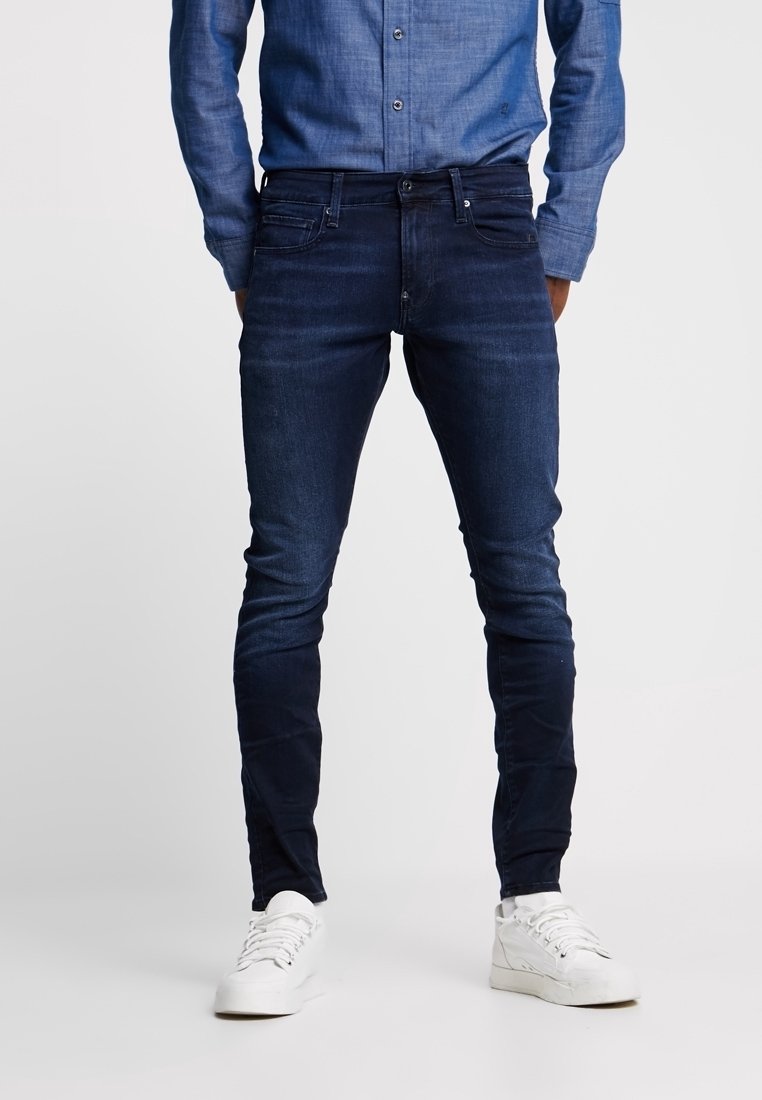 Man med mörkblå slim-fit jeans, blå skjorta med knappar och vita sneakers, stående mot en enkel ljus bakgrund.