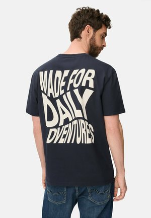Mężczyzna o ciemnych kręconych włosach i brodzie, ubrany w granatowy T-shirt z dużym białym napisem "MADE FOR DAILY ADVENTURES" na plecach, w połączeniu z niebieskimi dżinsami.