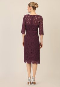 Abito di pizzo in viola scuro con maniche a metà, silhouette aderente, orlo scallop e cintura in raso. Indossato con sandali con tacco argento.