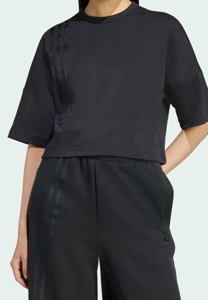 Osoba nosząca czarny krótki top na krótkie rękawy oraz dopasowane spodnie z wysokim stanem, ozdobione subtelnymi błyszczącymi paskami i logo na spodniach.