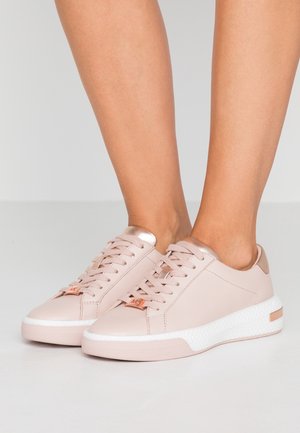 Femme portant des baskets basses rose pâle et or rose avec des semelles blanches texturées, debout sur un sol gris clair.