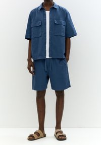 Chemise bleue à manches courtes avec boutons et deux poches poitrine, assortie à un short de la même couleur et des sandales marron, sur fond blanc.