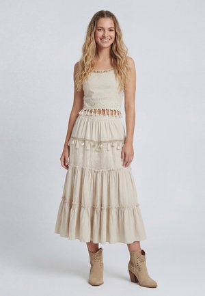 Młoda kobieta z długimi falującymi włosami, ubrana w beżowy bezrękawnik crop top z frędzlami i pasującą, wielowarstwową spódnicę oraz kowbojskie buty, uśmiechająca się.