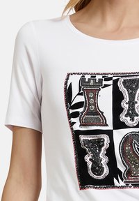 T-shirt bianca con una grafica quadrata che presenta motivi di pezzi degli scacchi in nero, rosso e con decorazioni argento. Maniche corte, scollatura rotonda.