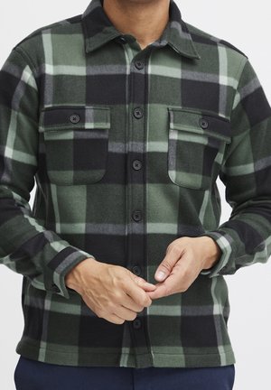 Homme portant une chemise à carreaux verte et noire avec poches poitrine, en train de boutonner, sur un fond uni.