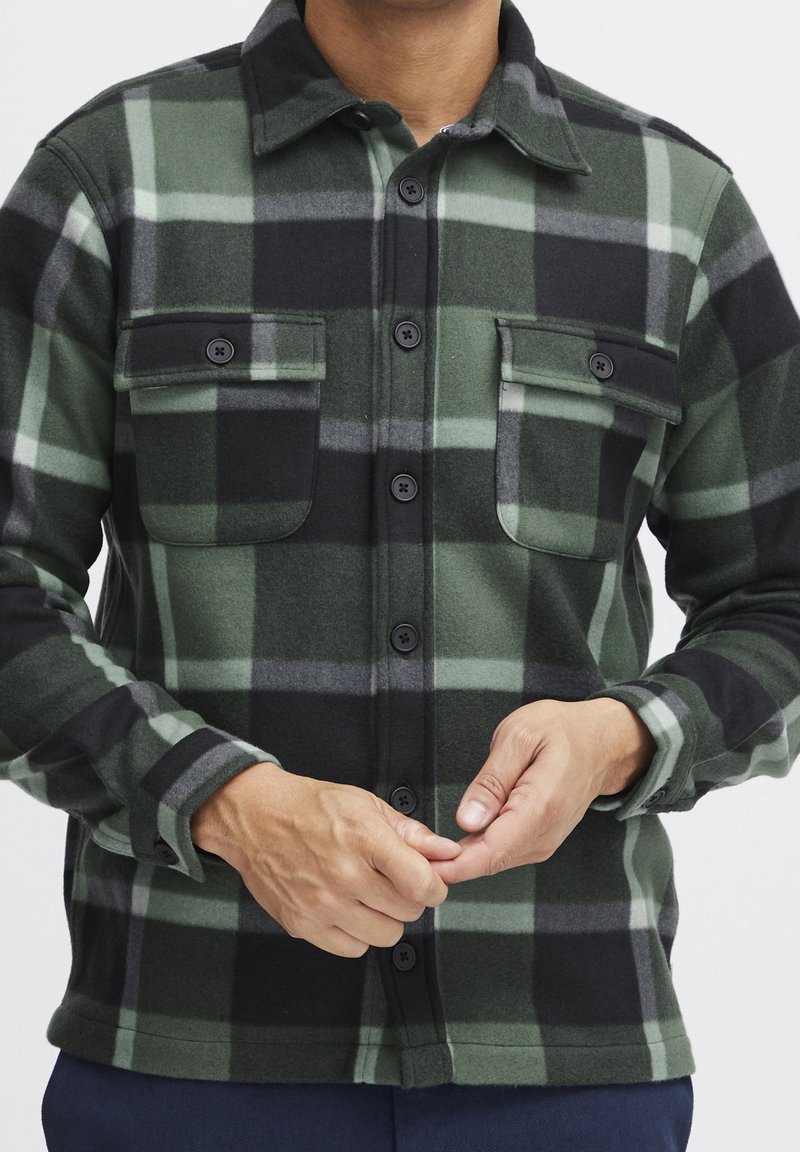 Homme portant une chemise à carreaux verte et noire avec poches poitrine, en train de boutonner, sur un fond uni.
