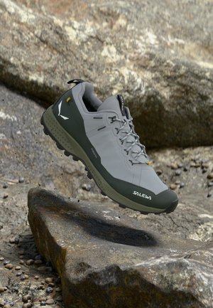 Világosszürke és zöld Salewa túracipő masszív talppal, fűzőkkel és logóval, sziklás terep felett elhelyezve.