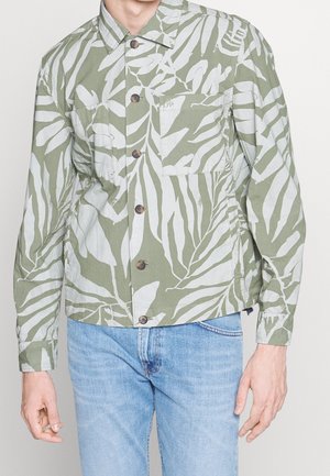 Homme portant une chemise verte à boutons avec un motif de feuilles blanches et un jean bleu clair, debout devant un fond uni.