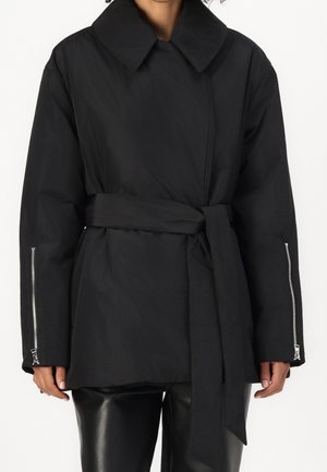 Femme portant un manteau noir ceinturé avec un large col et des fermetures éclair aux manches longues, associé à un pantalon en cuir noir.