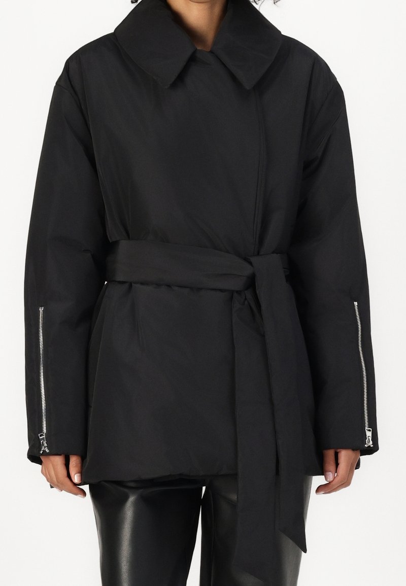 Femme portant un manteau noir ceinturé avec un large col et des fermetures éclair aux manches longues, associé à un pantalon en cuir noir.