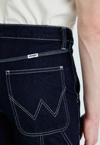 Dark blue denim jeans featuring a Wrangler label, contrasting white stitching, and a distinctive pocket design with a zigzag pattern.