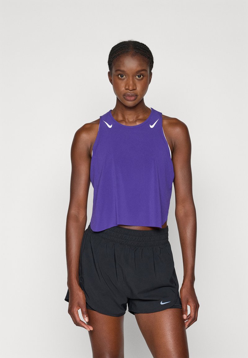 Fialová športová cropped tank top s bielymi akcentmi, doplnená čiernymi behacími šortkami so malým logom. Hladký materiál, voľný strih.