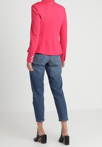 Blazer rose ajusté avec un design à basque, épaules structurées et poignets boutonnés, associé à un jean bleu taille haute et des talons noirs.