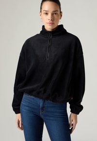 Μαύρο πουλόβερ από φλις με ψηλό γιακά και κλείσιμο με φερμουάρ στη μέση. Διαθέτει ριμπ μανσέτες και κοντό τελείωμα, συνδυασμένο με μπλε τζιν.