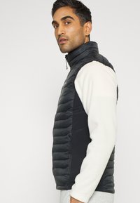 Gilet trapuntato nero con colletto alto, tasche con zip laterali e pannelli a contrasto. Indossato sopra un pile di colore chiaro.
