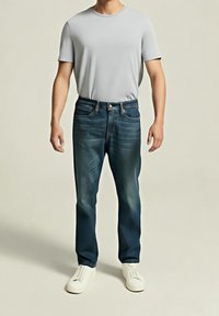 Homme portant une chemise à manches courtes gris clair, un jean bleu foncé et des chaussures blanches sans lacets sur un fond uni.