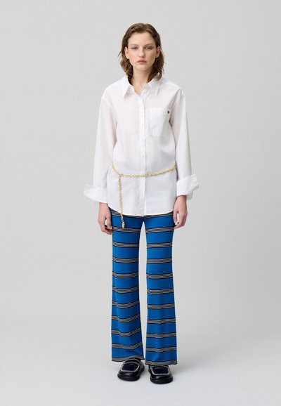 Camicia bianca a maniche lunghe con taschino sul petto, abbinata a pantaloni larghi a righe blu. Una cintura a catena dorata evidenzia il punto vita. Mocassini neri completano il look.