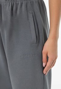 Sivé tepláky s elastickým pásom, bočnými vreckami a embosovaným logom. Tkanina je mäkká a hladká na dotyk.
