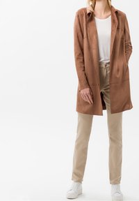 Chaqueta larga de ante marrón con cuello, bolsillos frontales, combinada con pantalones beiges y zapatillas blancas. Textura suave con un corte relajado.