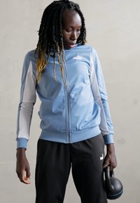 Svetlomodrá športová bunda s bielymi akcentmi, so zipsom vpredu a rebrovanými manžetami. Čierne nohavice. Drží čierny kettlebell.