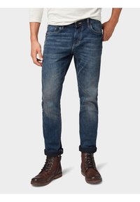 Jeans de mezclilla azules para hombre con ajuste slim, textura deslavada, diseño de cinco bolsillos y dobladillos enrollados, combinados con botas marrones de cordones.