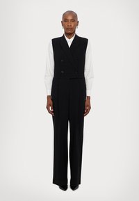 Jumpsuit negro a medida con un diseño de chaleco cruzado, pantalones de pierna ancha y una textura suave. Llevado sobre una camisa blanca con cuello.