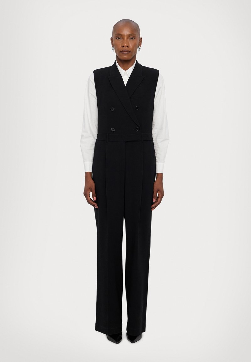 Jumpsuit negro a medida con un diseño de chaleco cruzado, pantalones de pierna ancha y una textura suave. Llevado sobre una camisa blanca con cuello.