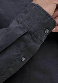 Manga de camisa de lino gris con cierre de botón, costura prominente y una superficie de tela texturizada. Se pueden ver dos botones oscuros.