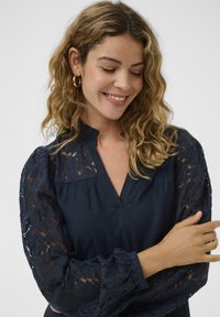 Blouse bleu marine avec manches bouffantes en dentelle et encolure en V. Présente un tissu texturé et des poignets froncés, ajoutant du détail au design.