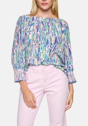 Frau trägt eine Bluse mit mehrfarbigem abstraktem Muster, Rüschenmanschetten und hellrosa hoch taillierte Hose, steht mit entspannten Händen.