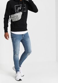 Felpa nera con logo bianco, borsa a tracolla grigia chiara, jeans skinny strappati blu e sneakers sportive bianche. Outfit casual.