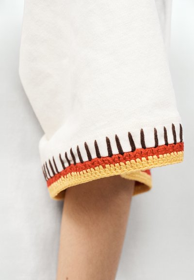 Biely bavlnený rukáv s farebnou háčkovanou obšívkou. Lem má oranžové, žlté a tmavohnedé akcenty v cikcak vzore.