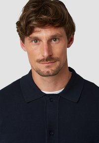 Marineblauwe polo met een kraag, voorzien van een knoopsluiting en een getextureerde stof. Model heeft halflang haar.