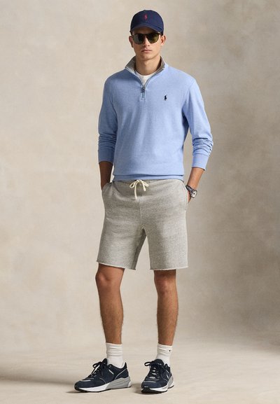 Férfi, aki sötétkék sapkát, napszemüveget, világoskék negyed cipzáras pulóvert, szürke húzózsinóros rövidnadrágot, fehér zoknit és sötétkék sportcipőt visel, semleges háttér előtt áll.