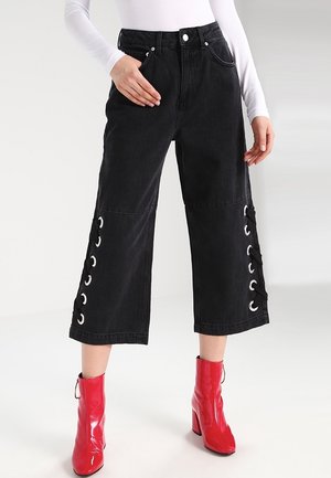 Pantalones culotte de mezclilla negra con detalles de ojal en los laterales, cintura alta y pierna ancha. Combinados con botas de tobillo rojas brillantes.