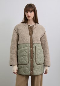 Chaqueta forrada de lana con parte superior beige, parte inferior acolchada en verde oliva y detalles en marrón. Cuenta con botones a presión y tela texturizada.