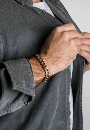 Pulsera de cadena de oro en una muñeca, con eslabones de acabado pulido. Llevada sobre una camisa de pana gris con las mangas arremangadas.