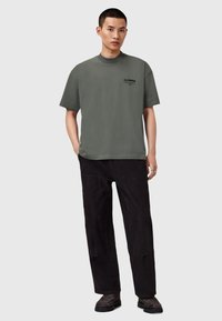 Camiseta de color verde oliva oscuro con cuello redondo y mangas cortas, combinada con pantalones de pana negros y zapatos negros texturizados.