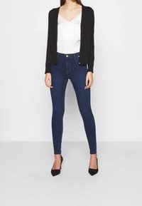 Svart cardigan, vit satin-topp, mörkblå tajta jeans och svarta spetsiga klackar; slank design, figurnära silhuett och mjuk textur.