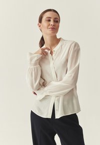 Blouse blanche à manches longues avec boutons sur le devant, coupe décontractée, poignets smockés et détail de bouton doré au col. Associée à un pantalon foncé.