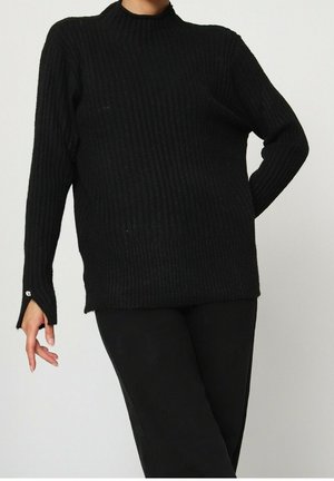 Pull long noir à côtes avec col montant et fentes latérales, avec des accents de boutons en acier inoxydable sur les poignets. Texture douce et coupe ample.